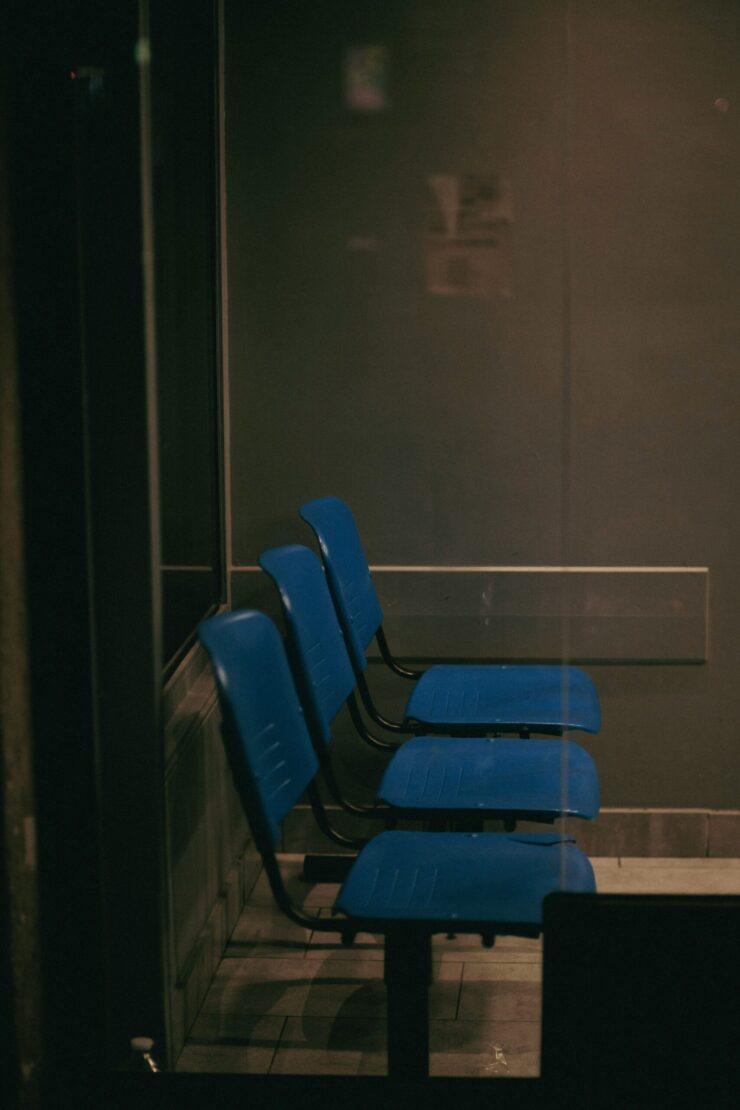 Vulnerabilidad humana: Imagen que muestra una fila de tres sillas de plástico azul en una sala de espera oscura y solitaria, vista a través de un cristal que refleja una atmósfera fría y burocrática.