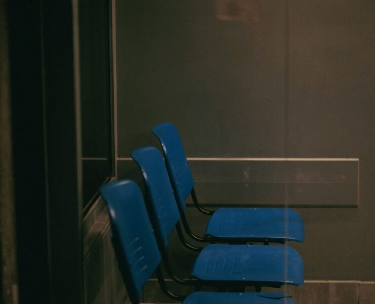 Vulnerabilidad humana: Imagen que muestra una fila de tres sillas de plástico azul en una sala de espera oscura y solitaria, vista a través de un cristal que refleja una atmósfera fría y burocrática.