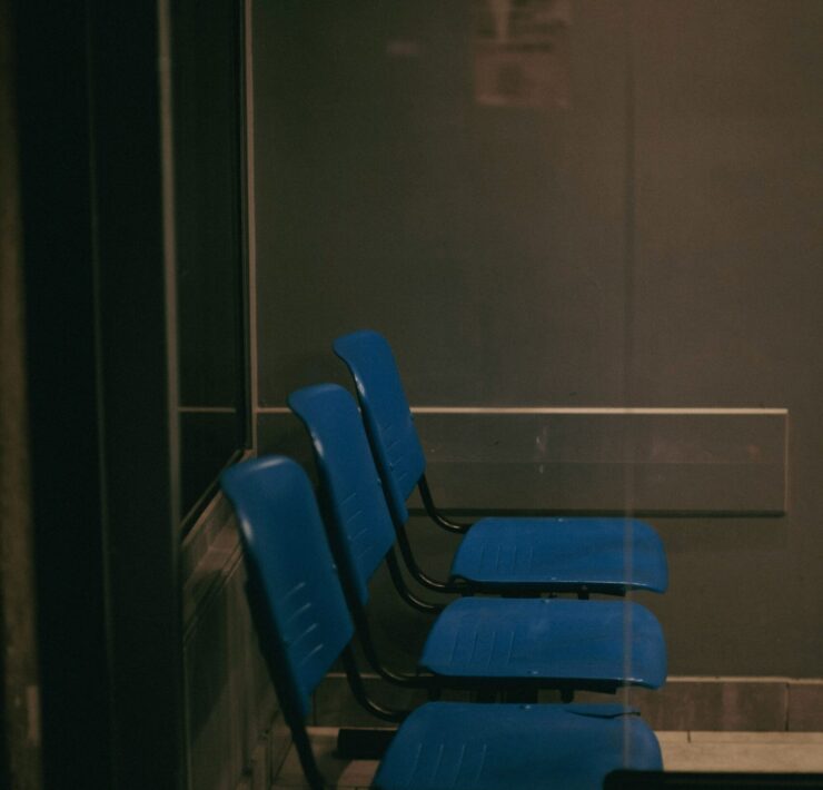 Vulnerabilidad humana: Imagen que muestra una fila de tres sillas de plástico azul en una sala de espera oscura y solitaria, vista a través de un cristal que refleja una atmósfera fría y burocrática.