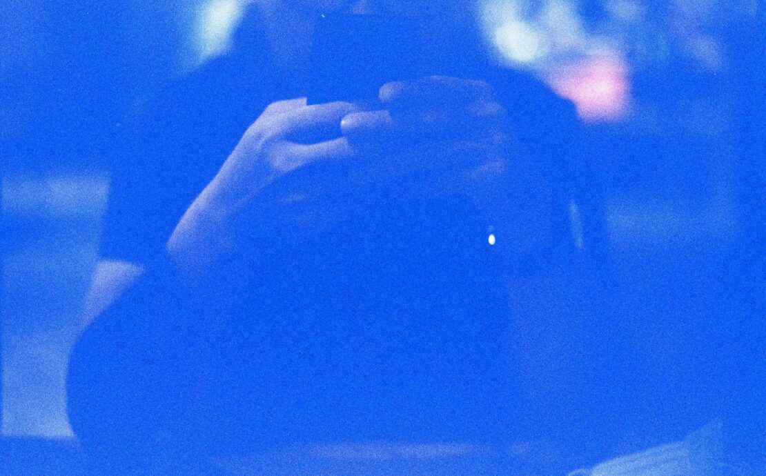 Identidad digital: retrato abstracto en tonos azules saturados de una silueta humana difusa frente a un dispositivo móvil. El fuerte grano fotográfico y el resplandor de la pantalla crean una atmósfera eléctrica donde los rasgos físicos desaparecen, dejando solo la forma del sujeto interactuando con la tecnología.