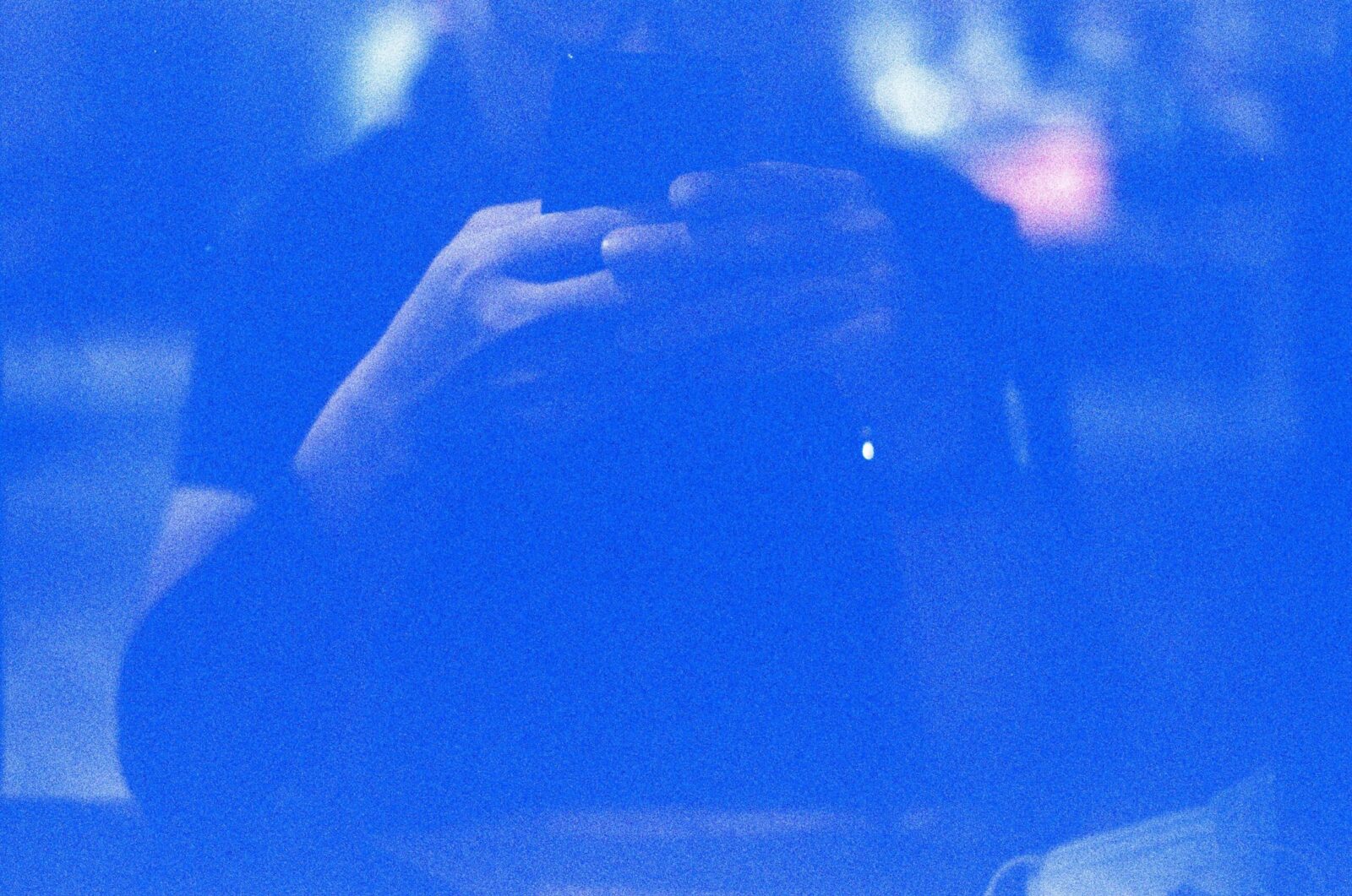 Identidad digital: retrato abstracto en tonos azules saturados de una silueta humana difusa frente a un dispositivo móvil. El fuerte grano fotográfico y el resplandor de la pantalla crean una atmósfera eléctrica donde los rasgos físicos desaparecen, dejando solo la forma del sujeto interactuando con la tecnología.