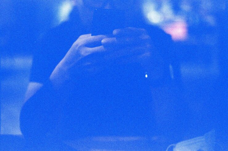 Identidad digital: retrato abstracto en tonos azules saturados de una silueta humana difusa frente a un dispositivo móvil. El fuerte grano fotográfico y el resplandor de la pantalla crean una atmósfera eléctrica donde los rasgos físicos desaparecen, dejando solo la forma del sujeto interactuando con la tecnología.