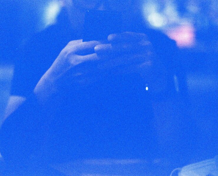 Identidad digital: retrato abstracto en tonos azules saturados de una silueta humana difusa frente a un dispositivo móvil. El fuerte grano fotográfico y el resplandor de la pantalla crean una atmósfera eléctrica donde los rasgos físicos desaparecen, dejando solo la forma del sujeto interactuando con la tecnología.