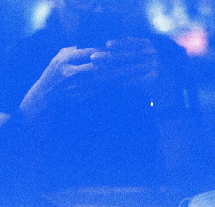 Identidad digital: retrato abstracto en tonos azules saturados de una silueta humana difusa frente a un dispositivo móvil. El fuerte grano fotográfico y el resplandor de la pantalla crean una atmósfera eléctrica donde los rasgos físicos desaparecen, dejando solo la forma del sujeto interactuando con la tecnología.