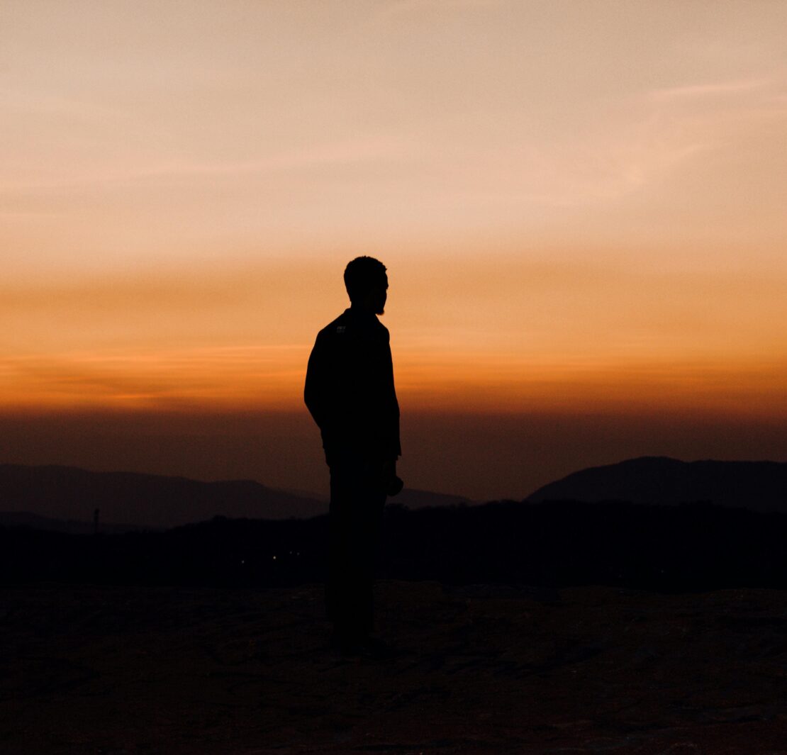 Silueta oscura de un hombre de perfil, de pie sobre una cima rocosa, contemplando un vasto horizonte de montañas bajo un cielo degradado en tonos naranja intenso y ocre al atardecer. La imagen captura una atmósfera de soledad y nostalgia, funcionando como una representación visual del amor inalcanzable.
