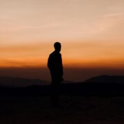 Silueta oscura de un hombre de perfil, de pie sobre una cima rocosa, contemplando un vasto horizonte de montañas bajo un cielo degradado en tonos naranja intenso y ocre al atardecer. La imagen captura una atmósfera de soledad y nostalgia, funcionando como una representación visual del amor inalcanzable.