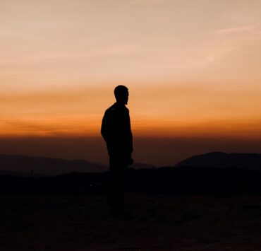 Silueta oscura de un hombre de perfil, de pie sobre una cima rocosa, contemplando un vasto horizonte de montañas bajo un cielo degradado en tonos naranja intenso y ocre al atardecer. La imagen captura una atmósfera de soledad y nostalgia, funcionando como una representación visual del amor inalcanzable.