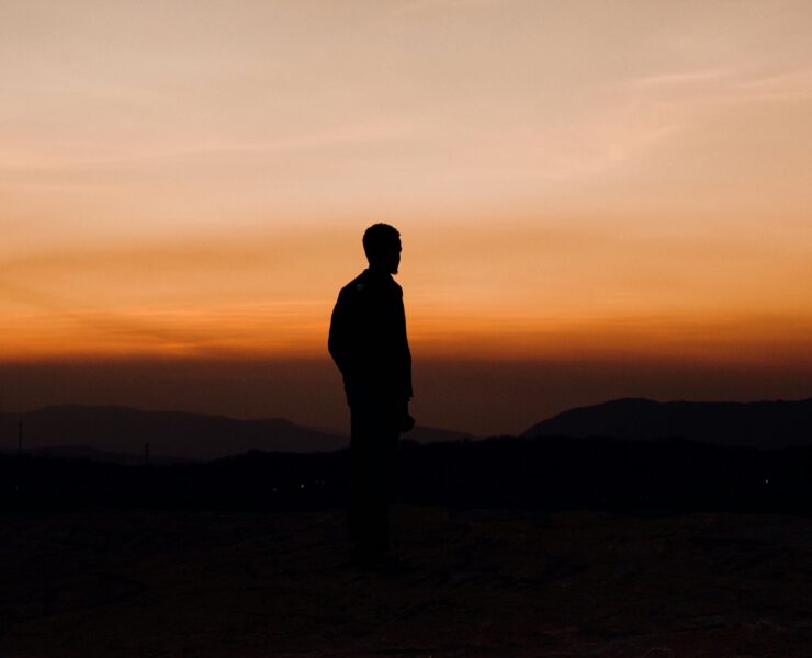 Silueta oscura de un hombre de perfil, de pie sobre una cima rocosa, contemplando un vasto horizonte de montañas bajo un cielo degradado en tonos naranja intenso y ocre al atardecer. La imagen captura una atmósfera de soledad y nostalgia, funcionando como una representación visual del amor inalcanzable.