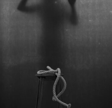 Herencia del dolor: fotografía artística en blanco y negro que muestra un taburete rústico con una cuerda de cáñamo enrollada en primer plano, mientras al fondo se proyecta la silueta sombría de una persona atrapada tras una tela translúcida, presionando sus manos en un gesto de angustia y súplica que simboliza la violencia psicológica y la fragilidad del alma ante el trauma.