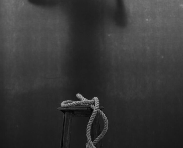 Herencia del dolor: fotografía artística en blanco y negro que muestra un taburete rústico con una cuerda de cáñamo enrollada en primer plano, mientras al fondo se proyecta la silueta sombría de una persona atrapada tras una tela translúcida, presionando sus manos en un gesto de angustia y súplica que simboliza la violencia psicológica y la fragilidad del alma ante el trauma.
