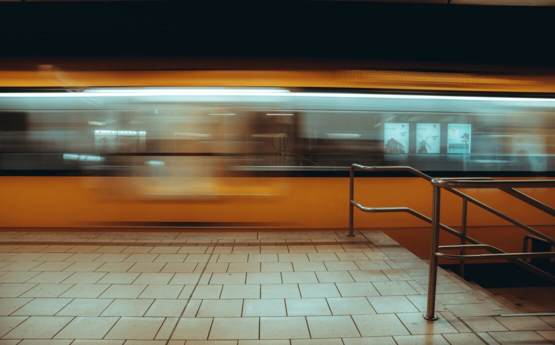 Multitud urbana: Imagen en plano general de una estación de tren subterránea; un tren amarillo se desplaza a gran velocidad creando una estela de movimiento frente a un andén de baldosas claras y una baranda metálica en primer plano.