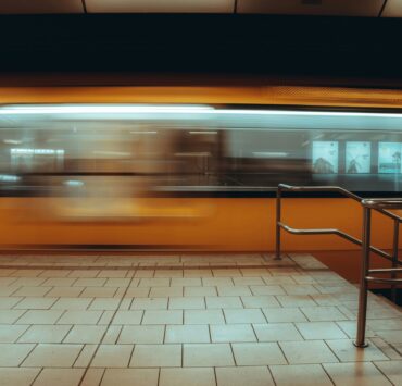 Multitud urbana: Imagen en plano general de una estación de tren subterránea; un tren amarillo se desplaza a gran velocidad creando una estela de movimiento frente a un andén de baldosas claras y una baranda metálica en primer plano.