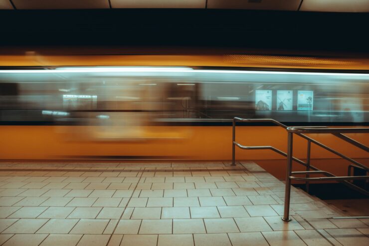 Multitud urbana: Imagen en plano general de una estación de tren subterránea; un tren amarillo se desplaza a gran velocidad creando una estela de movimiento frente a un andén de baldosas claras y una baranda metálica en primer plano.