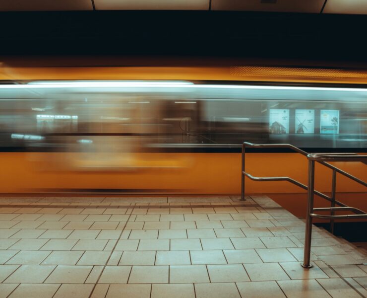 Multitud urbana: Imagen en plano general de una estación de tren subterránea; un tren amarillo se desplaza a gran velocidad creando una estela de movimiento frente a un andén de baldosas claras y una baranda metálica en primer plano.