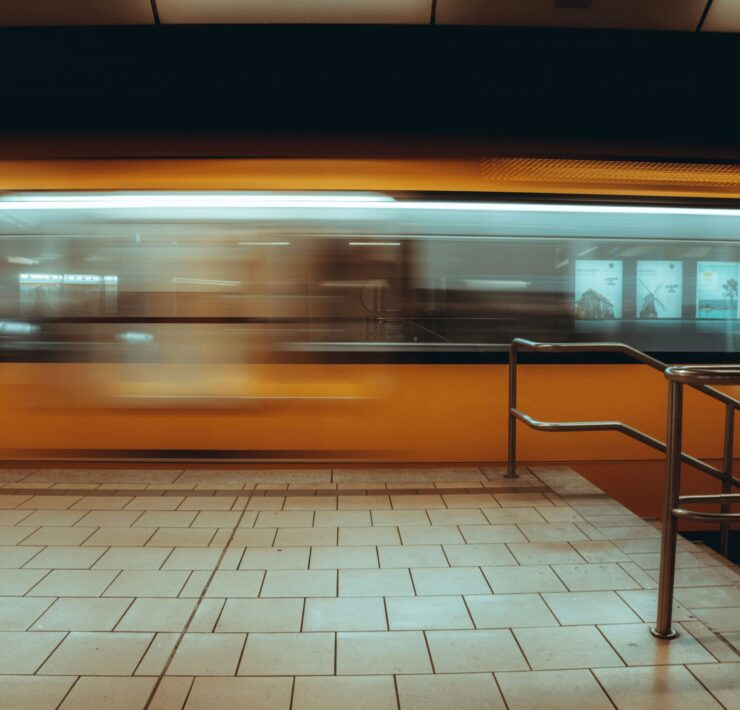 Multitud urbana: Imagen en plano general de una estación de tren subterránea; un tren amarillo se desplaza a gran velocidad creando una estela de movimiento frente a un andén de baldosas claras y una baranda metálica en primer plano.