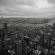 Fotografía panorámica aérea en blanco y negro de la ciudad de Seúl, Corea del Sur, dividida por el río Han y sus puentes. En primer plano se observa una densa acumulación de rascacielos y bloques de edificios bajo un cielo nublado, transmitiendo de forma visual el concepto de alienación social dentro de una megaurbe hiperproductiva.
