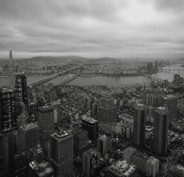 Fotografía panorámica aérea en blanco y negro de la ciudad de Seúl, Corea del Sur, dividida por el río Han y sus puentes. En primer plano se observa una densa acumulación de rascacielos y bloques de edificios bajo un cielo nublado, transmitiendo de forma visual el concepto de alienación social dentro de una megaurbe hiperproductiva.