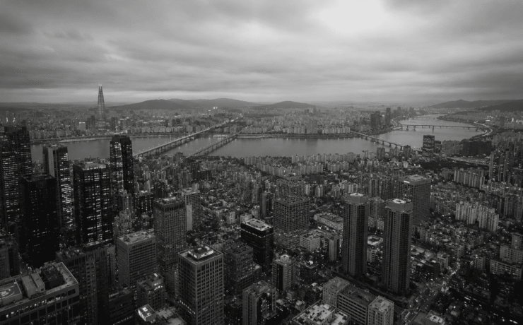 Fotografía panorámica aérea en blanco y negro de la ciudad de Seúl, Corea del Sur, dividida por el río Han y sus puentes. En primer plano se observa una densa acumulación de rascacielos y bloques de edificios bajo un cielo nublado, transmitiendo de forma visual el concepto de alienación social dentro de una megaurbe hiperproductiva.