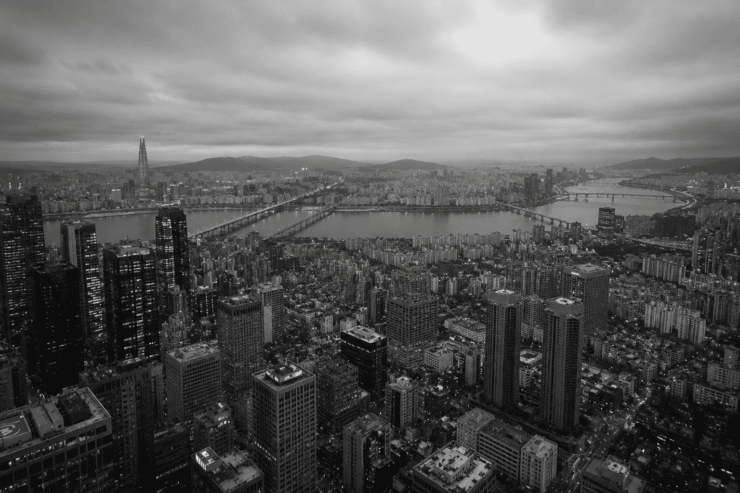 Fotografía panorámica aérea en blanco y negro de la ciudad de Seúl, Corea del Sur, dividida por el río Han y sus puentes. En primer plano se observa una densa acumulación de rascacielos y bloques de edificios bajo un cielo nublado, transmitiendo de forma visual el concepto de alienación social dentro de una megaurbe hiperproductiva.