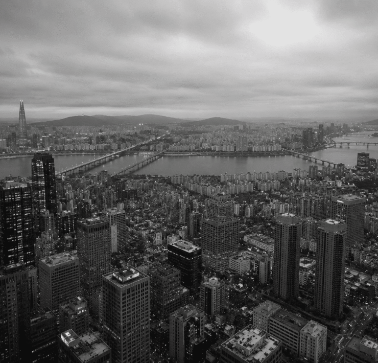 Fotografía panorámica aérea en blanco y negro de la ciudad de Seúl, Corea del Sur, dividida por el río Han y sus puentes. En primer plano se observa una densa acumulación de rascacielos y bloques de edificios bajo un cielo nublado, transmitiendo de forma visual el concepto de alienación social dentro de una megaurbe hiperproductiva.