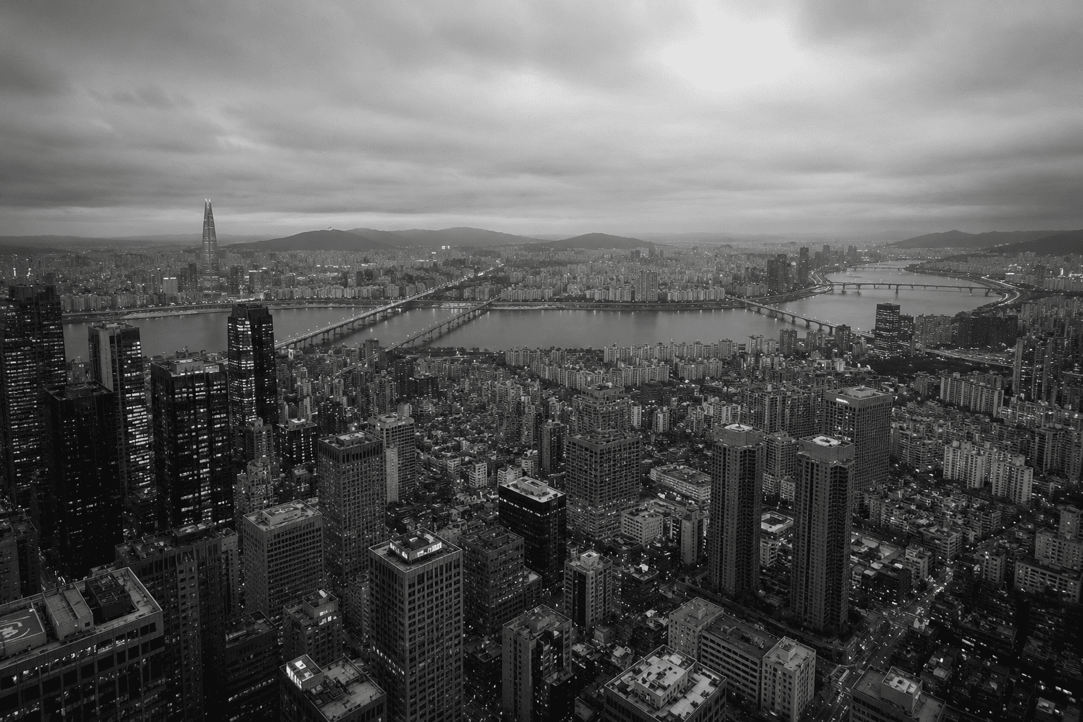 Fotografía panorámica aérea en blanco y negro de la ciudad de Seúl, Corea del Sur, dividida por el río Han y sus puentes. En primer plano se observa una densa acumulación de rascacielos y bloques de edificios bajo un cielo nublado, transmitiendo de forma visual el concepto de alienación social dentro de una megaurbe hiperproductiva.