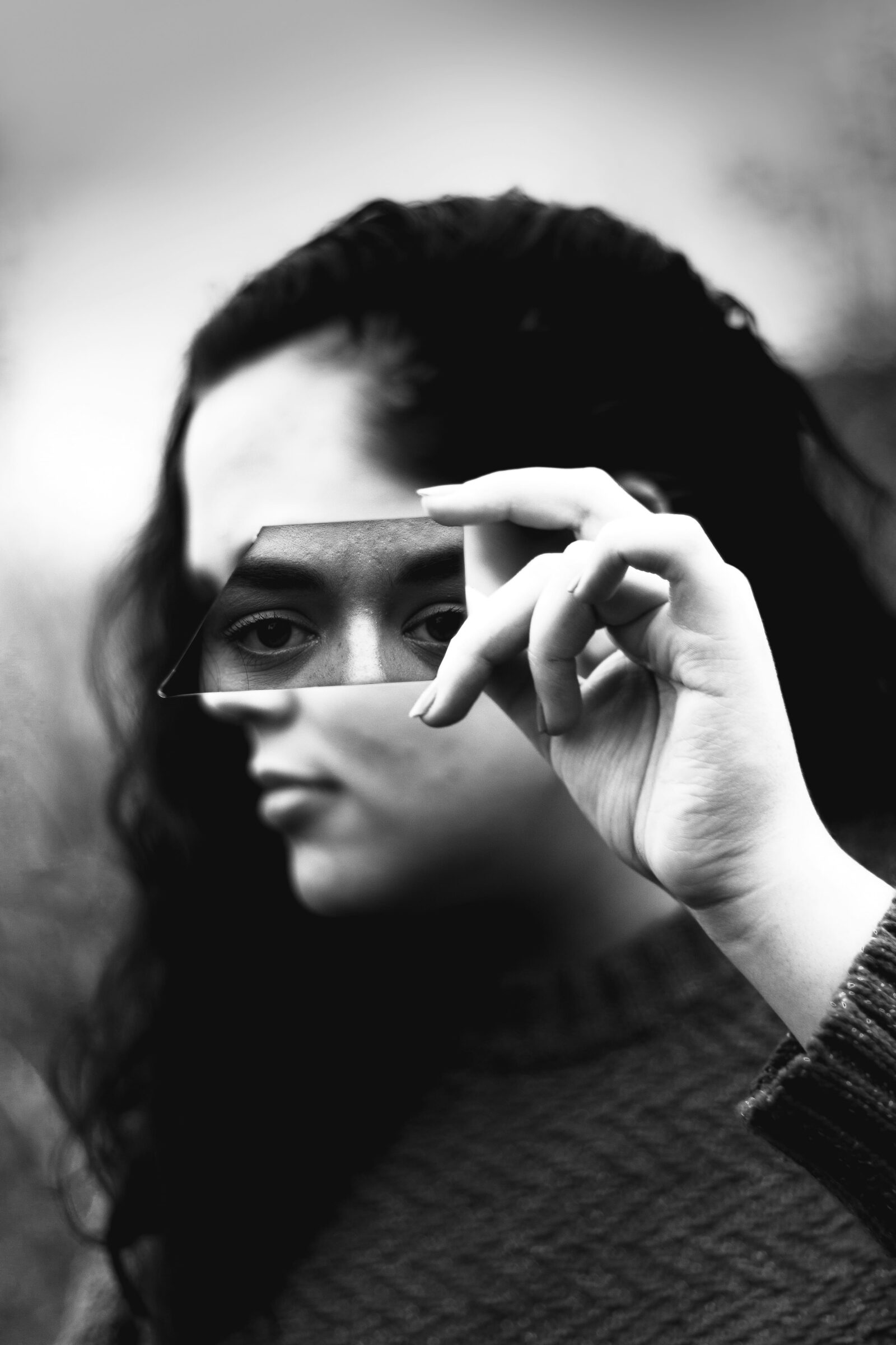 Fotografía en blanco y negro de primer plano. Una mujer sostiene horizontalmente con su mano derecha un fragmento alargado de vidrio roto justo a la altura de sus ojos. El trozo de espejo refleja de forma nítida su mirada fija y directa, transmitiendo un profundo cansancio emocional, mientras que el resto de su rostro y el fondo exterior quedan completamente borrosos y fuera de foco.