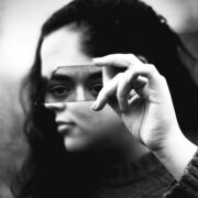 Fotografía en blanco y negro de primer plano. Una mujer sostiene horizontalmente con su mano derecha un fragmento alargado de vidrio roto justo a la altura de sus ojos. El trozo de espejo refleja de forma nítida su mirada fija y directa, transmitiendo un profundo cansancio emocional, mientras que el resto de su rostro y el fondo exterior quedan completamente borrosos y fuera de foco.