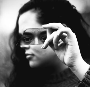 Fotografía en blanco y negro de primer plano. Una mujer sostiene horizontalmente con su mano derecha un fragmento alargado de vidrio roto justo a la altura de sus ojos. El trozo de espejo refleja de forma nítida su mirada fija y directa, transmitiendo un profundo cansancio emocional, mientras que el resto de su rostro y el fondo exterior quedan completamente borrosos y fuera de foco.