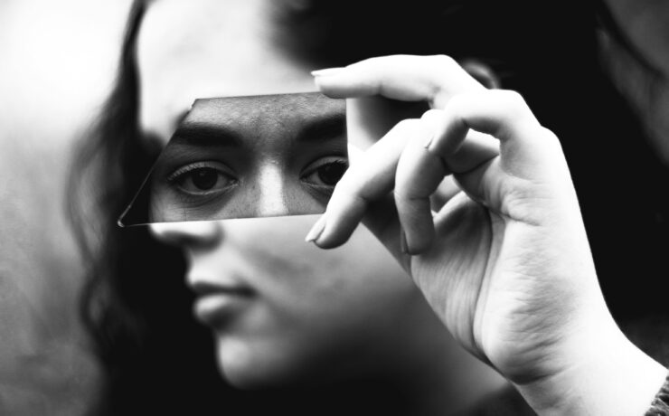 Fotografía en blanco y negro de primer plano. Una mujer sostiene horizontalmente con su mano derecha un fragmento alargado de vidrio roto justo a la altura de sus ojos. El trozo de espejo refleja de forma nítida su mirada fija y directa, transmitiendo un profundo cansancio emocional, mientras que el resto de su rostro y el fondo exterior quedan completamente borrosos y fuera de foco.
