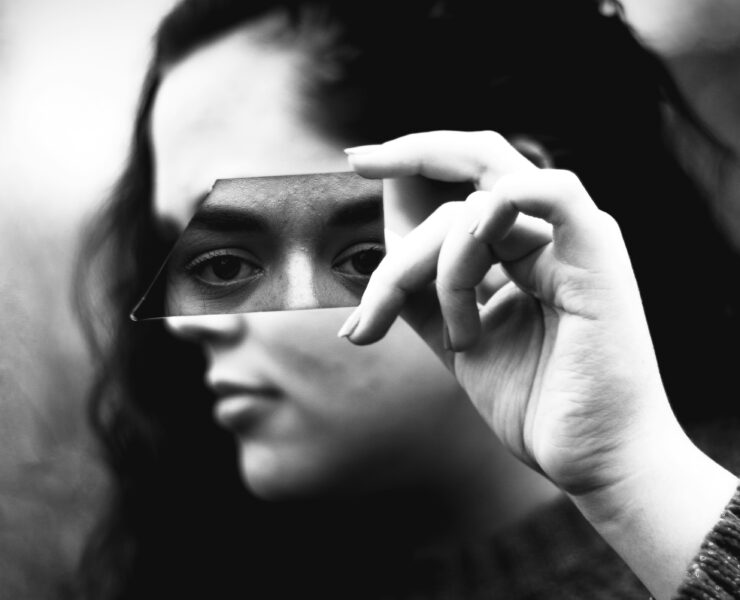 Fotografía en blanco y negro de primer plano. Una mujer sostiene horizontalmente con su mano derecha un fragmento alargado de vidrio roto justo a la altura de sus ojos. El trozo de espejo refleja de forma nítida su mirada fija y directa, transmitiendo un profundo cansancio emocional, mientras que el resto de su rostro y el fondo exterior quedan completamente borrosos y fuera de foco.